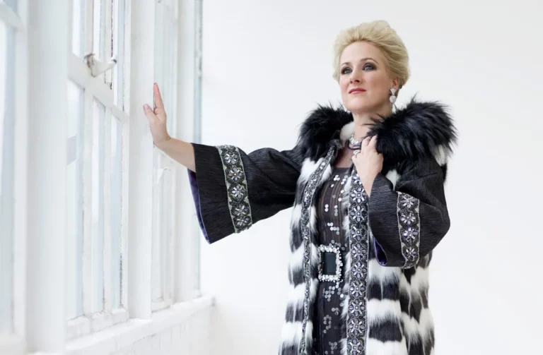 Soprano Diana Damrau in un elegante cappotto di pelliccia, in piedi vicino a una finestra, catturata in un momento di bellezza.